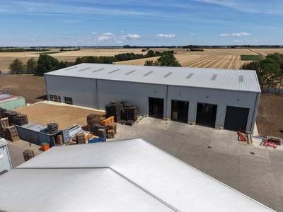 Drone view of the completed steel-framed building: front elevation with four roller-shutter bays opening onto a new concrete yard, open farmland beyond