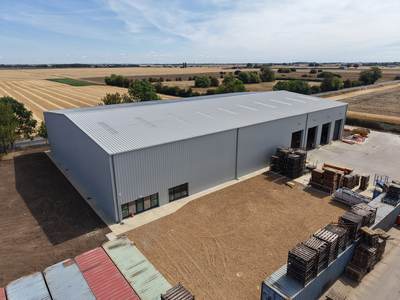 Drone view of the completed building from the corner, showing the clad side elevation with windows and the concrete yard