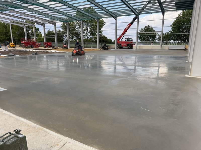 Operator power-floating a large concrete floor slab inside a steel-framed industrial building